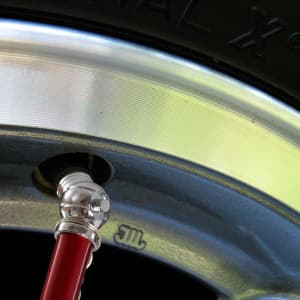 A close-up of a silver wheel rim and a red pressure gauge inserted into the tyre valve. The image features a metallic finish.