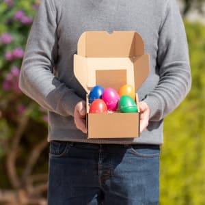 A person holding an open cardboard box containing colourful plastic eggs in various bright colours.