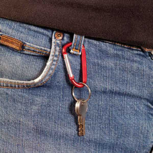 A red and silver carabiner attached to a person's jeans, holding a key. Features a logo on the carabiner.