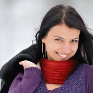 A woman wearing a red neck warmer smiles while holding a black jacket over her shoulder.
