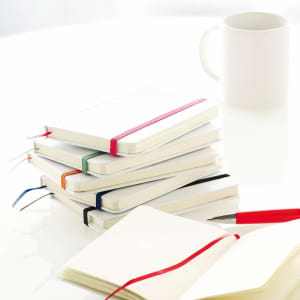 A stack of notepads in various colours with elastic bands, beside an empty mug and a red pen.