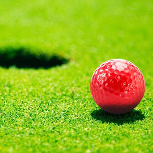 A red golf ball sits on lush green grass, near a golf hole. The ball features a dimpled surface and has a logo.