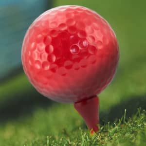 A red golf tee with a textured surface, placed on green grass. The background features a blurred golf club.