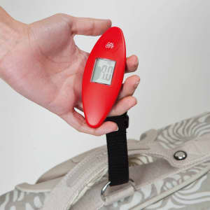 A red digital luggage scale held in a hand, displaying a weight reading, next to a patterned bag.