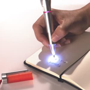 A silver and red pen with a built-in torch lights up while writing in a notebook, alongside an orange cap.