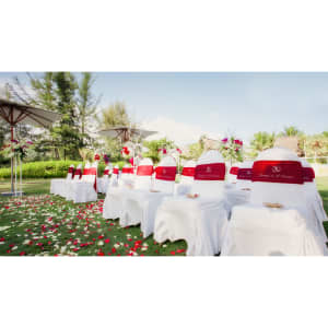 A row of white chairs adorned with red bands, set outdoors amidst flowers and greenery under umbrellas.