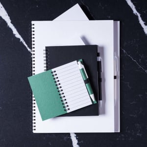 A collection of notebooks in green, black, and white, stacked on a dark marble surface, accompanied by a pen.