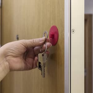 A hand holding keys is placing them onto a red keyring hook mounted on a wooden door.
