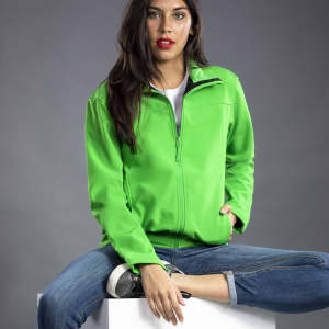 A bright green soft shell jacket with a zip, worn by a model seated on a white cube, featuring a logo.