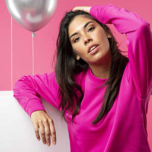 A woman in a bright pink sweatshirt poses with one hand on her head, set against a pink background with a silver balloon.