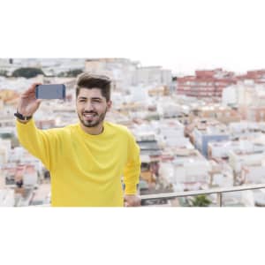 A man wearing a yellow sweatshirt takes a selfie against a cityscape background. The sweatshirt has a logo.