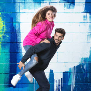 A pink technical jacket and a black jacket are worn by a woman and a man, respectively, against a colourful wall backdrop.
