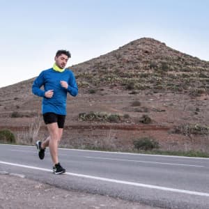 An adult male runner in a blue long-sleeve shirt and black shorts jogs along a road with hills in the background.