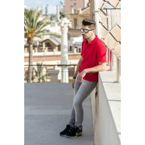 A red Bartel polo shirt worn by a man leaning against a wall, paired with grey pants and sneakers.