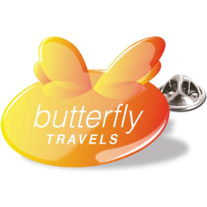 A vibrant orange lapel pin featuring butterfly wings and a logo for "butterfly TRAVELS." It has a metal backing.