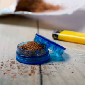 A blue grinder with finely ground material sits on a wooden surface beside a yellow lighter.