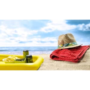 A yellow table holds snacks and drinks with a straw hat and a red towel on the sandy beach under a blue sky.
