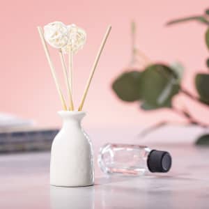 A white ceramic vase holds three flower-shaped reeds, accompanied by a clear glass bottle with a black lid.