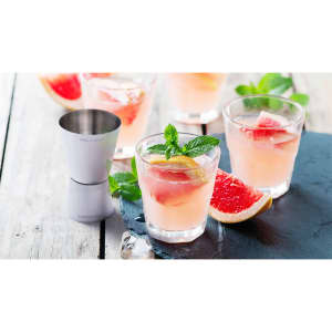 A stainless steel jigger with drinks featuring pink grapefruit, mint leaves, and slices of lemon in glasses.
