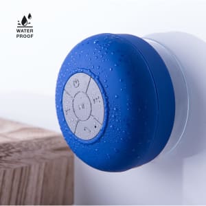 A blue waterproof speaker features a round design with buttons, resting against a white background next to wood.