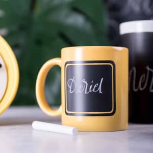 A yellow coffee mug with a chalkboard surface and a white chalk stick next to it, featuring a logo.