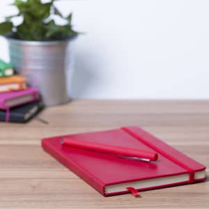 A red notebook with an elastic band and a matching pen, placed on a wooden desk alongside a plant.