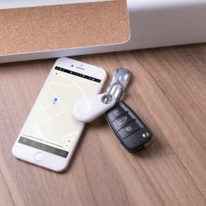 A white Krosly Tracker attached to car keys, placed next to a smartphone displaying a map on a wooden surface.