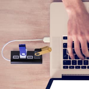 A black USB hub connected to a laptop, with blue and gold USB drives inserted. A hand reaches for the keyboard.