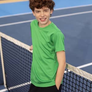 A bright green kids' T-shirt with a textured design, featuring a logo, worn by a smiling boy on a tennis court.