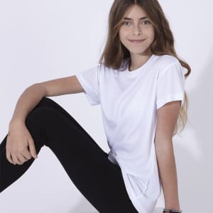 A white kids' T-shirt displayed on a girl seated with black leggings. The shirt features a logo.