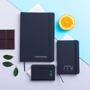 A set of black notebooks in various sizes with a logo, accompanied by orange slices, chocolate pieces, and green leaves.