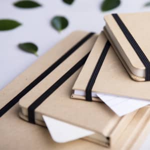A series of kraft paper notepads in varying sizes, featuring black elastic bands, arranged with green leaves in the background.
