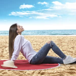 A woman relaxes on a red mat on the beach, wearing a white top and blue jeans, with a straw hat nearby.