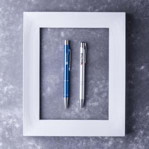 Two metal pens are placed inside a white frame on a textured grey background. One pen is blue and the other is silver.