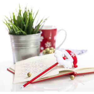 A festive red pen with a Santa hat on a notebook, with a decorative plant and a Christmas-themed mug in the background.