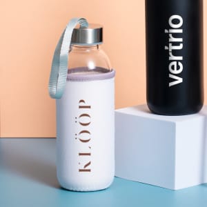 A drink bottle with a clear glass body, featuring a white protective sleeve and a silver lid, alongside a black bottle.