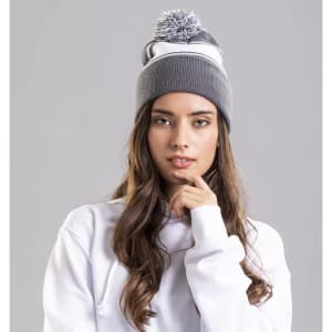 A grey beanie with a pom-pom, worn by a model with long hair, set against a neutral background.
