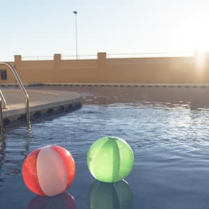 Two beach balls, one red and white and the other green, float in a pool under a sunny sky.