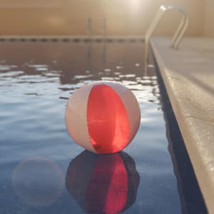 A beach ball with red and white panels floats on the water near a poolside.