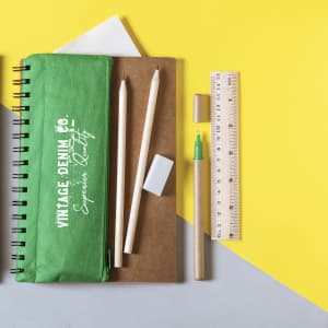 A green notebook with a logo, pencils, a ruler, an eraser, and a pen on a yellow and grey background.