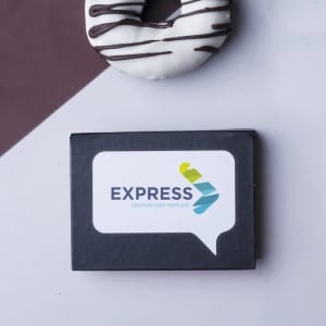 A black box with a logo and a sticky notepad inside, placed beside a chocolate-covered donut.