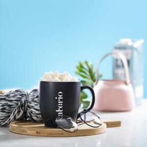 A black coffee mug filled with marshmallows, resting on a wooden tray, with sunglasses nearby and a pastel backdrop.
