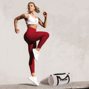 A fitness enthusiast in a white crop top and red leggings demonstrates a high-knee exercise beside a black and white branded duffle bag.