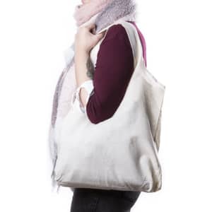 A cream tote bag is being held by a person wearing a scarf, showcasing its spacious design and simplicity.