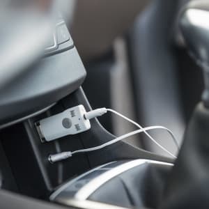 A white Bluetooth receiver is plugged into a car's audio port, with a headphone jack cable connected nearby.