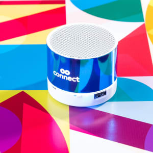 A small, round blue speaker with a white base, featuring a logo, placed on a colourful geometric background.