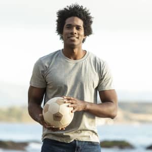 A man stands outdoors wearing a grey MC150 Adult Colour T-Shirt, holding a soccer ball with a scenic background.