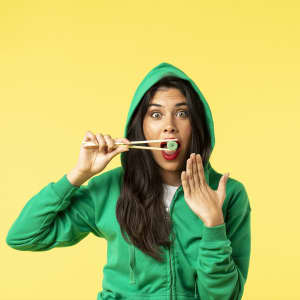 A woman in a green hooded sweatshirt holds sushi with chopsticks, displaying surprise against a yellow background.