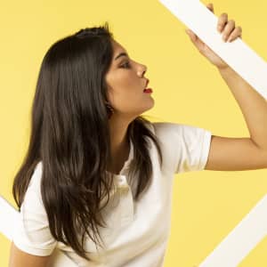 A woman with long dark hair poses in a white polo shirt against a yellow background. The shirt has a branding logo.