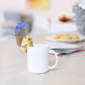 A white mug with a yellow duck planter and purple flowers, featuring a small logo on the side.